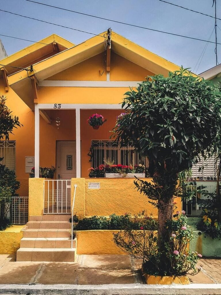 Fachada de una casa color amarillo con detalles en blanco y escaleras en la entrada y un árbol adornando en la banqueta